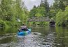 man in a blue sit-in kayak on the Black River. A bridge over the river can be seen in the distance