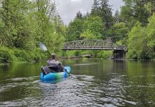 man in a blue sit-in kayak on the Black River. A bridge over the river can be seen in the distance