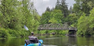 man in a blue sit-in kayak on the Black River. A bridge over the river can be seen in the distance