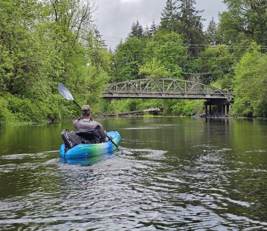man in a blue sit-in kayak on the Black River. A bridge over the river can be seen in the distance